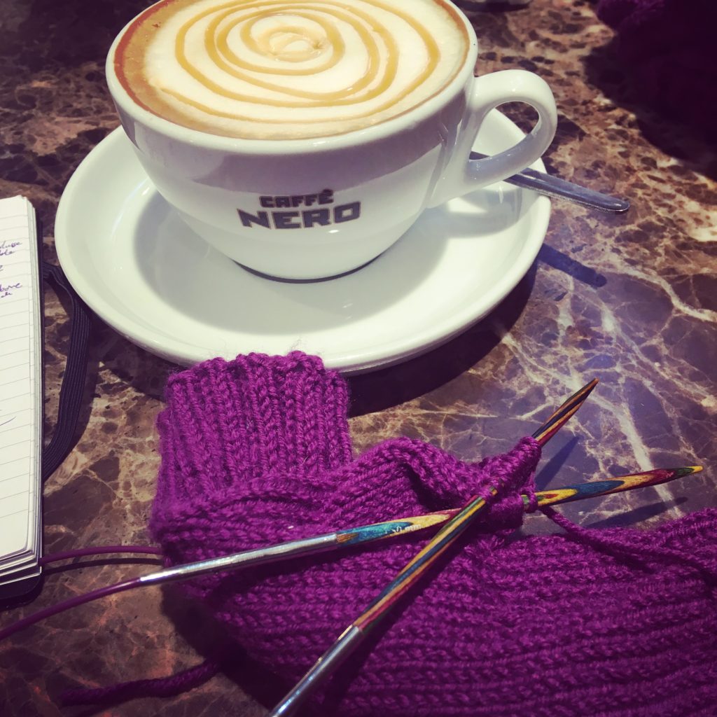 In the foreground there is a purple mitten with knitting needles through part of them. There is a coffee cup in the background