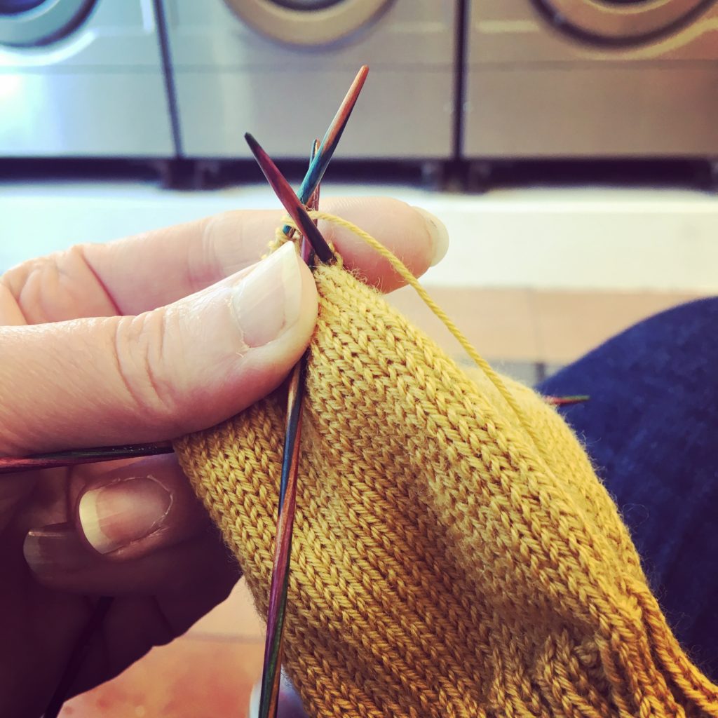 Hand holding some mustard yellow knitting on double-point needles. There's a row of washing machines in the background.